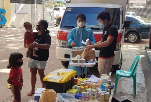 Healthcare workers wearing face masks distribute medicines and supplies from a table beside a van while assisting local residents, including a woman holding a child and a young girl waiting nearby, in an outdoor community setting.